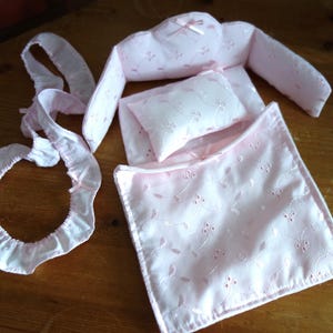 May include: A baby bedding set in soft pink, featuring a pillow, bumper pads, and a fitted sheet. The set is adorned with delicate floral embroidery and small bows. The pieces are arranged on a wooden surface, showcasing the details of the nursery decor.