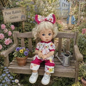May include: A doll with blonde hair and blue eyes wearing a white top with a colourful floral and rabbit pattern, matching cropped trousers with a pink hem, and white shoes. A large matching floral bow is worn in the hair, and the doll sits on a bench.