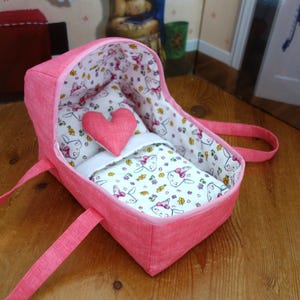May include: A pink fabric doll carrier with two handles, featuring a white interior lining patterned with small cartoon rabbits and flowers. The carrier includes a matching white pillow and blanket, topped with a small, solid pink heart-shaped cushion.