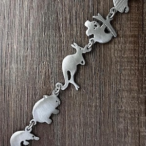 May include: A silver bracelet with charms depicting Australian wildlife, such as a kangaroo, koala, and echidna. The bracelet is made of a shiny metal and is shown on a wooden background. The detailed charms are linked together.