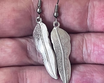 May include: A pair of silver feather earrings with a detailed textured design. The earrings are on a silver hook.