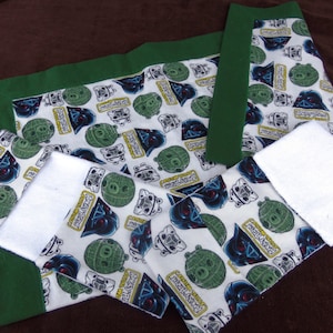 May include: A white baby blanket with a green border and a repeating pattern of Darth Vader and a green sphere. The blanket is folded in half and has a white terry cloth backing.
