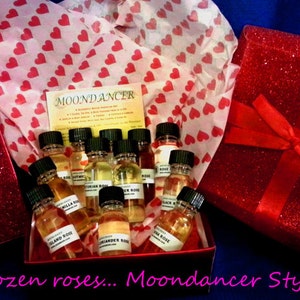 May include: A red gift box with a red ribbon and the words "For You" on the front. The box is filled with small bottles of essential oil, each with a different label. The bottles are arranged in a circle, and the box is surrounded by pink tissue paper with a heart pattern. The text "MOONDANCER" is visible on a card in the center of the image. The text "a dozen roses... Moondancer Style!" is visible at the bottom of the image.