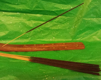 May include: A wooden incense holder with a crescent moon design and a single black incense stick burning on top. A bundle of incense sticks is lying on a green surface.