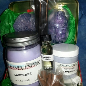 May include: A lavender-themed gift set in a green and gold tin box. The set includes a 16 oz. soy candle, body lotion, essential oil, incense sticks, and a lavender-scented soap. The products are all labeled with the brand name "Moondancer".