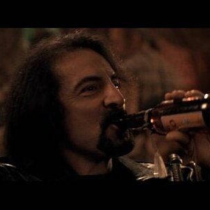 May include: A man with long dark hair drinks from a brown glass bottle with a white label. The bottle is held in his right hand.