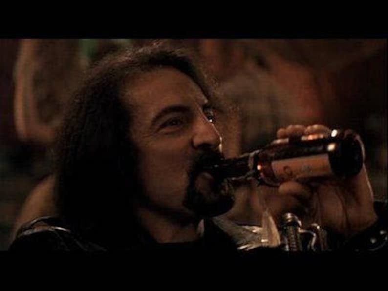 May include: A man with long dark hair drinks from a brown glass bottle with a white label. The bottle is held in his right hand.