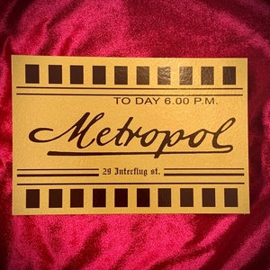 May include: A vintage-style sign with a gold background and black lettering. The sign reads "TO DAY 6.00 P.M. Metropol 29 Interflug st."