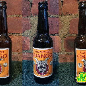May include: Three brown glass bottles of Cerveza Chango beer. Each bottle has an orange label with a red and white illustration of a snarling creature with horns. The label also includes the text "Cerveza Chango" in a white font.