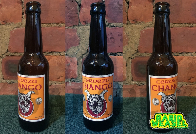 May include: Three brown glass bottles of Cerveza Chango beer. Each bottle has an orange label with a red and white illustration of a snarling creature with horns. The label also includes the text "Cerveza Chango" in a white font.