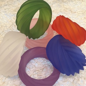 May include: A collection of colorful, abstract glass art pieces in shades of red, blue, green, pink, purple, and white. The shapes include a leaf, twisted forms, and oval rings. The pieces are arranged on a textured, off-white surface.