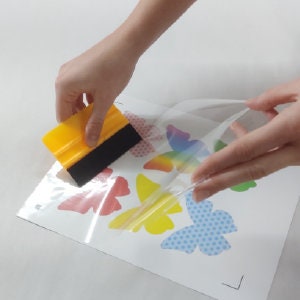 May include: A clear plastic sheet with a variety of colorful butterfly designs printed on it. A person is using a yellow and black scraper to smooth out the sheet.