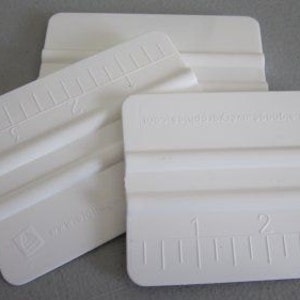 Puede incluir: Tres rasquetas de plástico blanco con una regla en la parte inferior. Las rasquetas se utilizan para aplicar calcomanías y pegatinas de vinilo.