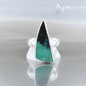Pode incluir: Um anel de prata com uma gema triangular verde e preta engastada no centro. O anel está em um expositor branco. O texto "Apsaras V" é visível no fundo.