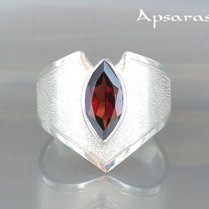 Puede incluir: Un anillo de plata con una piedra preciosa roja engastada en un diseño en forma de V. El anillo tiene una superficie texturizada.