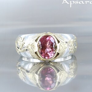 Puede incluir: Un anillo de plata y oro con una piedra preciosa ovalada rosa engastada en el centro. El anillo presenta intrincados diseños florales en la banda.