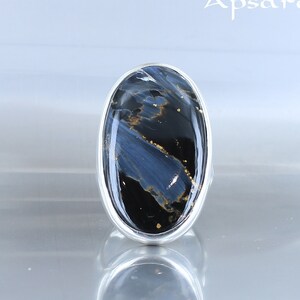 Puede incluir: Un anillo de plata con una gran piedra ovalada azul y negra con motas doradas. La piedra tiene un patr&oacute;n de remolino.