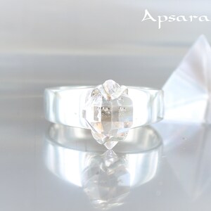 Puede incluir: Un anillo de plata con una piedra preciosa transparente, facetada, en forma de diamante engastada en el centro. El anillo está sobre una superficie blanca.