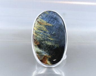 Peut inclure: Une bague en argent avec une pierre en œil de tigre bleu, or et noir. La pierre est de forme ovale et sertie dans une bélière en argent.