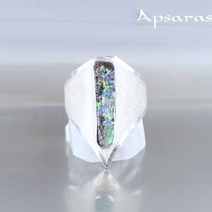 K&ouml;nnte beinhalten: Ein silberner Ring mit einer strukturierten Band und einem rechteckigen Opal-Stein, der in der Mitte eingesetzt ist. Der Opal hat eine Vielzahl von Farben, darunter Gr&uuml;n, Blau und Lila.
