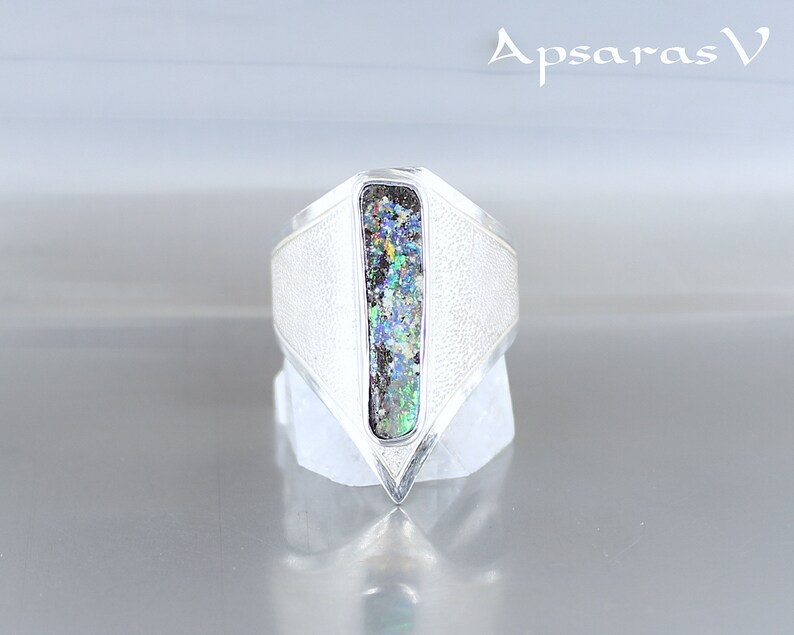 K&ouml;nnte beinhalten: Ein silberner Ring mit einer strukturierten Band und einem rechteckigen Opal-Stein, der in der Mitte eingesetzt ist. Der Opal hat eine Vielzahl von Farben, darunter Gr&uuml;n, Blau und Lila.