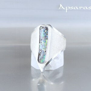 K&ouml;nnte beinhalten: Ein silberner Ring mit einem dreieckigen Design und einem rechteckigen, schillernden Opal-Stein, der in der Mitte eingesetzt ist. Der Ring befindet sich auf einem wei&szlig;en St&auml;nder.