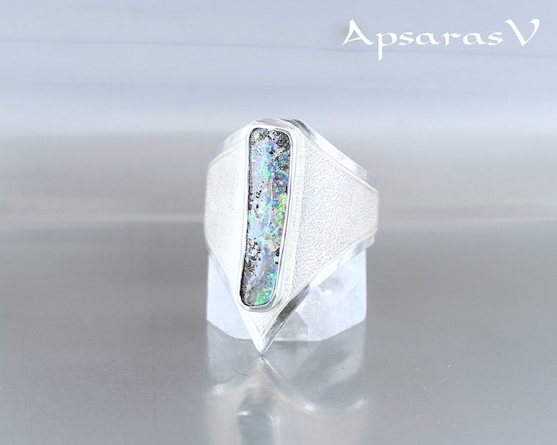 K&ouml;nnte beinhalten: Ein silberner Ring mit einem dreieckigen Design und einem rechteckigen, schillernden Opal-Stein, der in der Mitte eingesetzt ist. Der Ring befindet sich auf einem wei&szlig;en St&auml;nder.