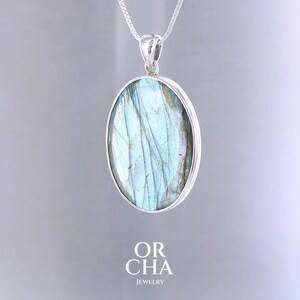 Peut inclure: Un collier pendentif en argent avec une pierre ovale de labradorite. La pierre présente des nuances de bleu et de gris avec des veines naturelles. Le pendentif est suspendu à une fine chaîne en argent. Le texte "OR CHA JEWELRY" est en bas.