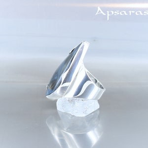 Peut inclure: Une bague en argent avec une pierre pr&eacute;cieuse bleue et grise sertie dans un design angulaire unique. La bague est pr&eacute;sent&eacute;e sur un rocher blanc.