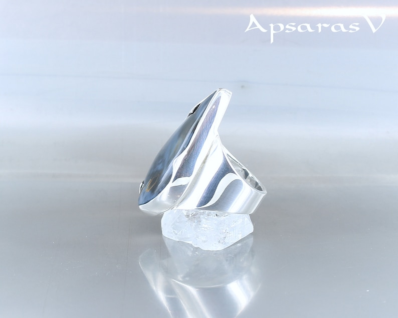 Peut inclure: Une bague en argent avec une pierre pr&eacute;cieuse bleue et grise sertie dans un design angulaire unique. La bague est pr&eacute;sent&eacute;e sur un rocher blanc.