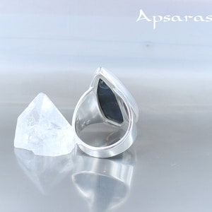 Peut inclure: Une bague en argent avec une pierre pr&eacute;cieuse noire sertie en forme de larme. La bague est pos&eacute;e sur une surface blanche avec un petit cristal transparent &agrave; c&ocirc;t&eacute;.