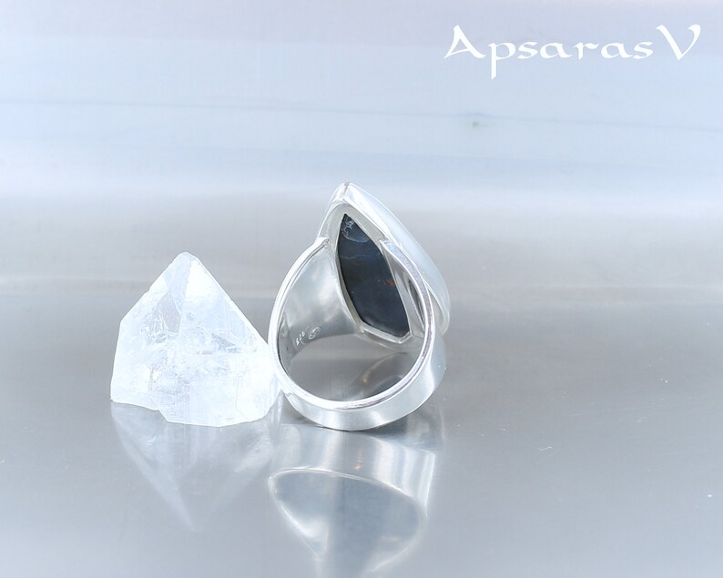 Peut inclure: Une bague en argent avec une pierre pr&eacute;cieuse noire sertie en forme de larme. La bague est pos&eacute;e sur une surface blanche avec un petit cristal transparent &agrave; c&ocirc;t&eacute;.