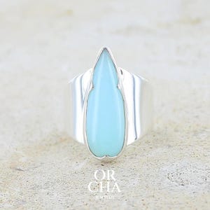 May include: A polished silver ring featuring a central, smooth, teardrop-shaped light blue stone set in a pointed bezel. The band is wide and reflective, with the text OR CHA JEWELRY printed in white letters at the bottom of the image frame.