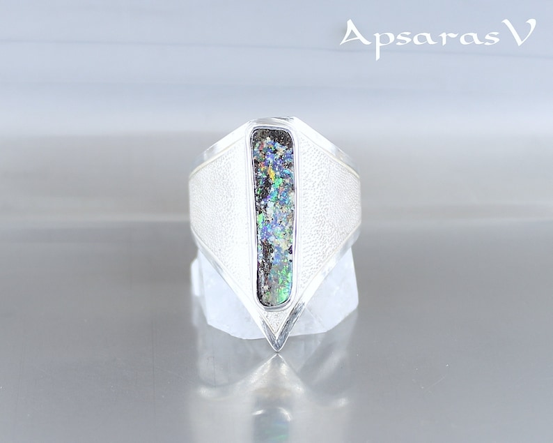 K&ouml;nnte beinhalten: Ein silberner Ring mit einer strukturierten Band und einem rechteckigen Opal-Stein, der in der Mitte eingesetzt ist. Der Ring ist in Form eines spitzen Dreiecks.
