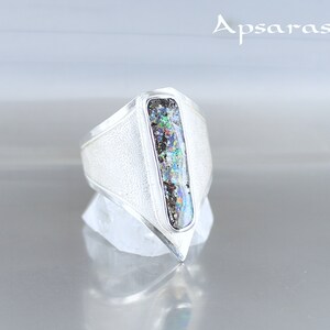 K&ouml;nnte beinhalten: Ein silberner Ring mit einem rechteckigen, schillernden Opal-Stein, der in einem spitz zulaufenden, geometrischen Design gefasst ist.