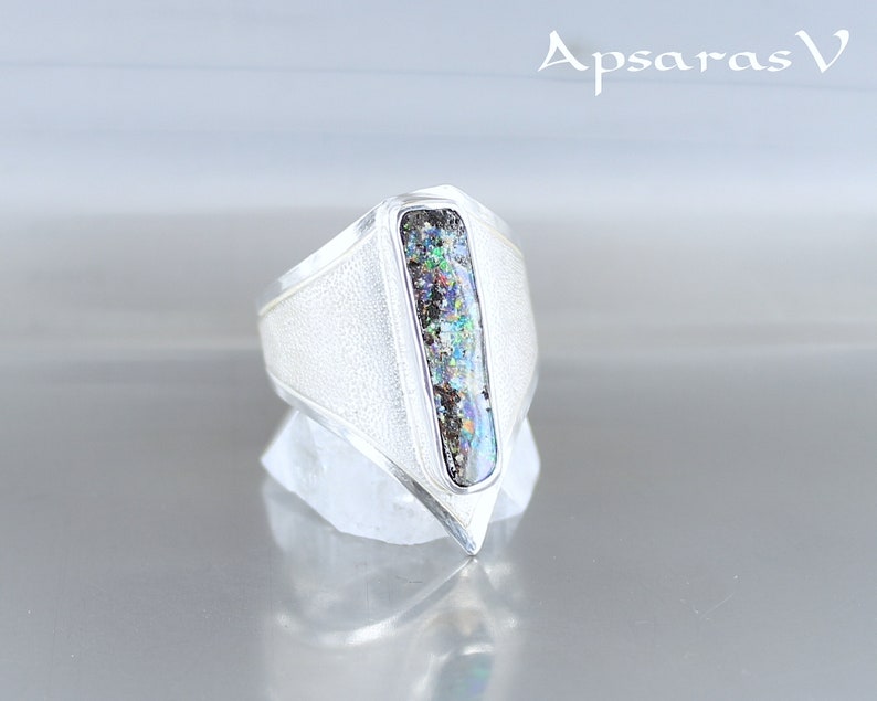 K&ouml;nnte beinhalten: Ein silberner Ring mit einem rechteckigen, schillernden Opal-Stein, der in einem spitz zulaufenden, geometrischen Design gefasst ist.