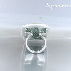 May include: A silver ring with a green and gold stone set in a square bezel. The ring is on a white surface.