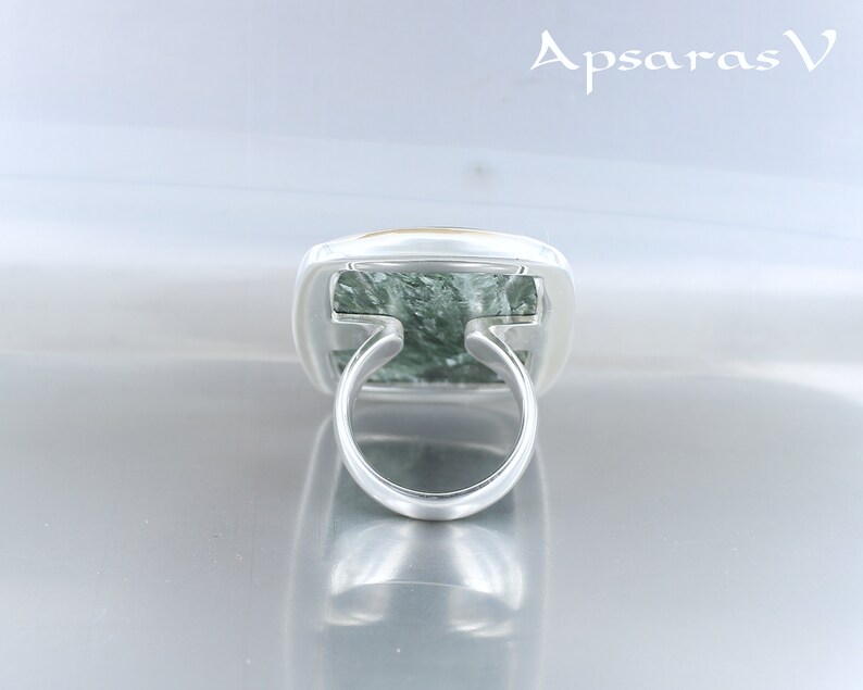 May include: A silver ring with a green and gold stone set in a square bezel. The ring is on a white surface.