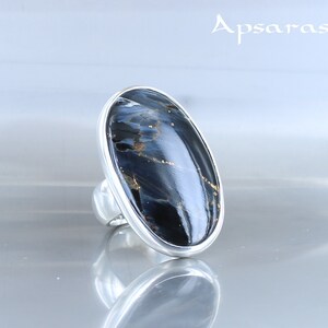 Puede incluir: Un anillo de plata con una gran piedra ovalada de ojo de tigre azul. La piedra tiene una base azul oscuro con vetas doradas.