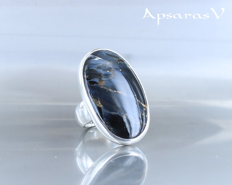 Puede incluir: Un anillo de plata con una gran piedra ovalada de ojo de tigre azul. La piedra tiene una base azul oscuro con vetas doradas.
