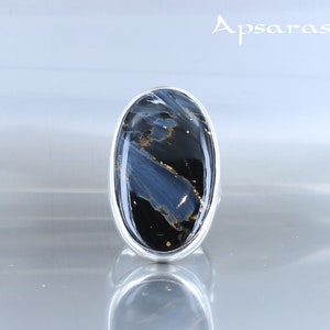 Puede incluir: Un anillo de plata con una gran piedra ovalada azul y negra con motas doradas. La piedra tiene un patr&oacute;n de remolino.