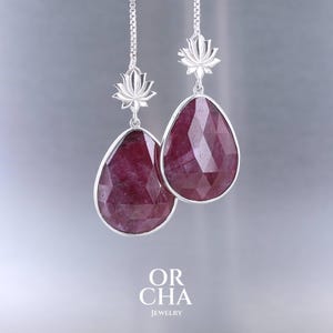 Puede incluir: Pendientes de plata con gemas facetadas en forma de lágrima de color rojo intenso. Cada pendiente tiene un diseño de flor de loto en la parte superior y el texto "OR CHA JEWELRY" en la parte inferior.