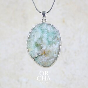 Op de afbeelding: Een hangerketting met een onregelmatige, ovaalvormige druzy-steen met lichtgroene en witte kristallijne texturen, gevat in een gepolijste zilverkleurige metalen rand en hangend aan een fijne zilverkleurige ketting. Tekst: OR CHA JEWELRY.