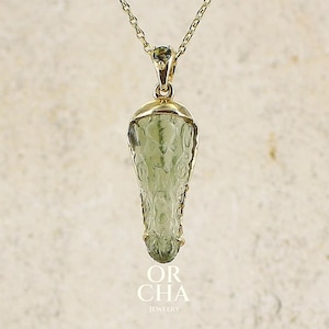 Peut inclure: Un collier pendentif en or avec une pierre vert pâle translucide et texturée en forme de goutte, sertie dans une monture en or avec une petite pierre verte sur la bélière. Le texte OR CHA JEWELRY est imprimé en blanc sous le pendentif.