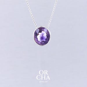 May include: A silver necklace featuring a large, oval-cut amethyst pendant. The purple gemstone is set in a silver bezel and hangs from a delicate silver chain. The necklace is displayed against a light gray background. The brand name "OR CHA JEWELRY" is at the bottom.