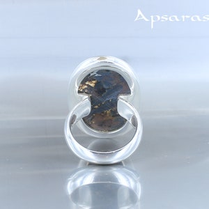 Puede incluir: Un anillo de plata con una piedra preciosa marr&oacute;n oscuro y dorada engastada en un engaste de bisel. El anillo se muestra desde la parte posterior, revelando la banda de plata lisa.