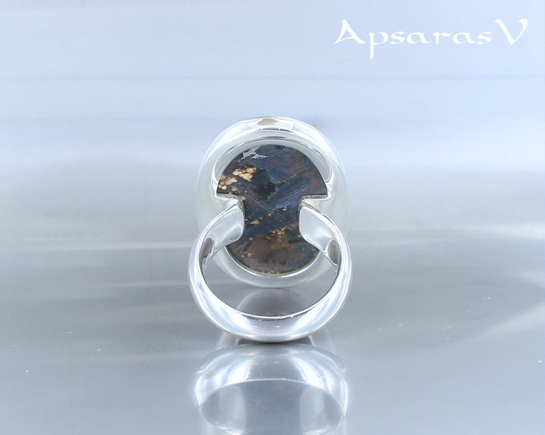 Puede incluir: Un anillo de plata con una piedra preciosa marr&oacute;n oscuro y dorada engastada en un engaste de bisel. El anillo se muestra desde la parte posterior, revelando la banda de plata lisa.