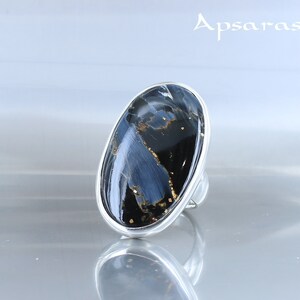Puede incluir: Un anillo de plata con una gran piedra ovalada azul y negra con motas doradas. El anillo est&aacute; sobre un fondo blanco.