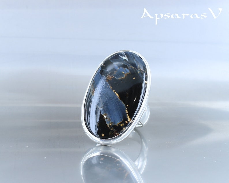 Puede incluir: Un anillo de plata con una gran piedra ovalada azul y negra con motas doradas. El anillo est&aacute; sobre un fondo blanco.
