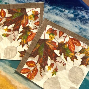 May include: Two white paper napkins with a brown border. The napkins feature an autumnal design with red, orange, yellow, and green leaves.
